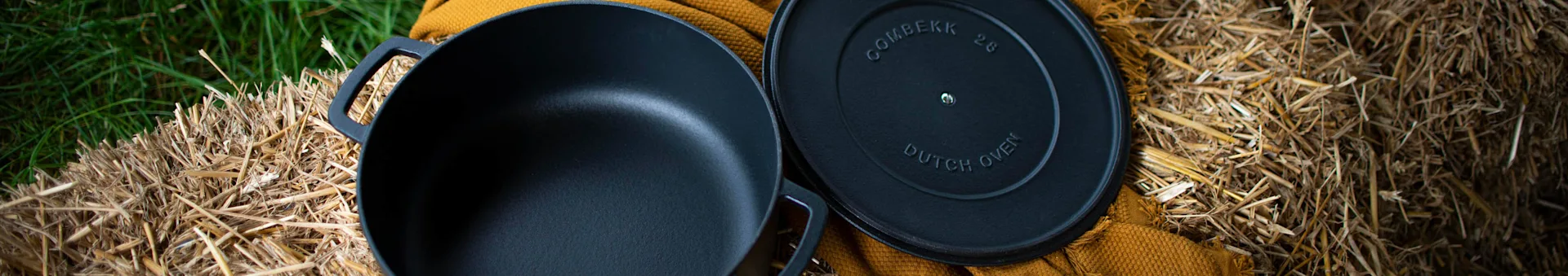 A black cast iron Combekk 2.0 Dutch oven with its lid, on hay bales and green grass, with a yellow fabric.