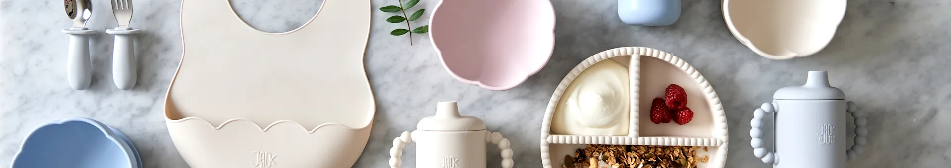 Flat lay of baby feeding items: cream bib, pink bowl, divided plate with yogurt and fruit, and two sippy cups, on marble.