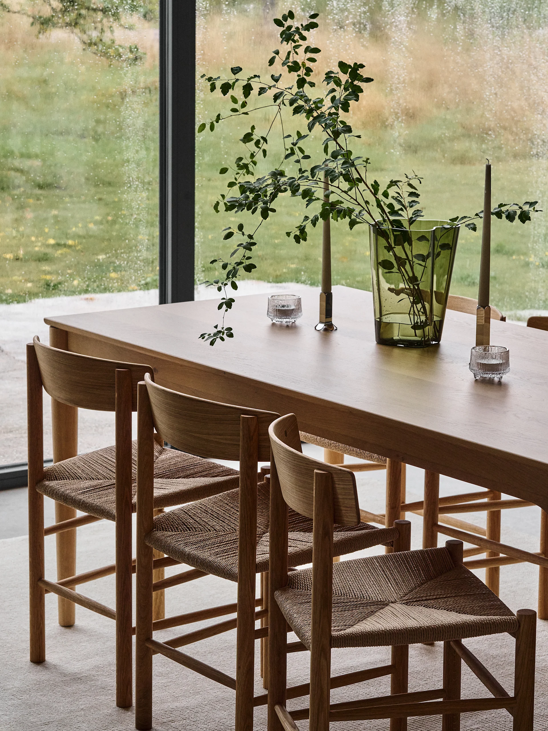 Dining room with light wood table, woven chairs, a green vase with branches, and candles against a rainy window.