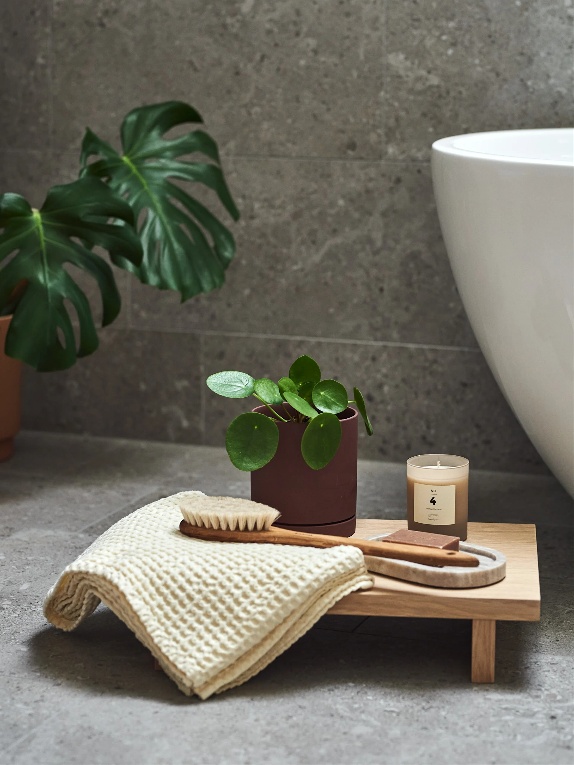 A Pilea plant, lemon verbena candle, body brush, soaps, and waffle towel on a wooden tray in a tiled bathroom.
