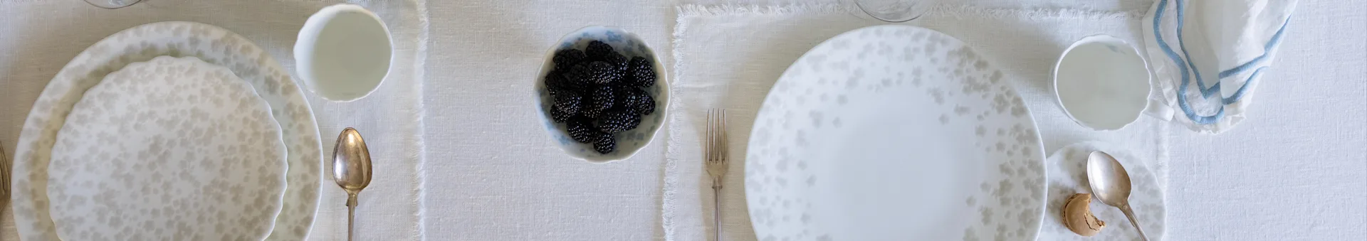 Overhead view of a dining table set for two with patterned white plates, silverware, blackberries, and a blue-trimmed napkin.