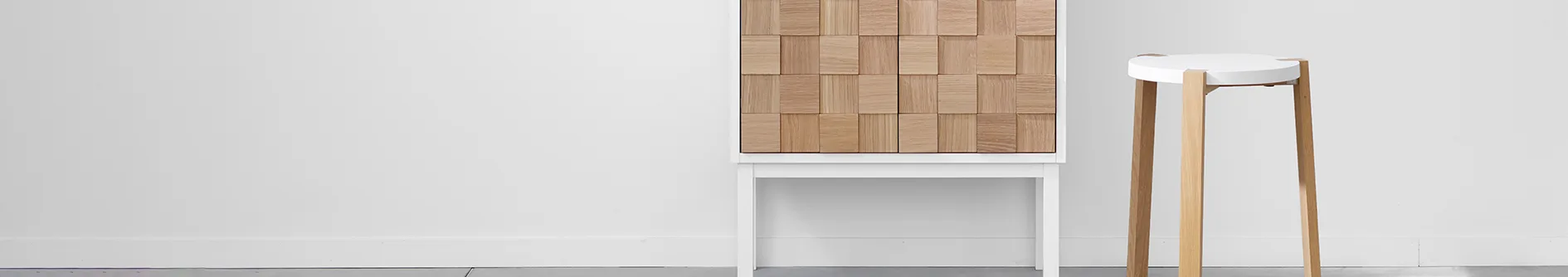 A white cabinet with a textured wooden front and a tall white stool with wooden legs against a white wall.