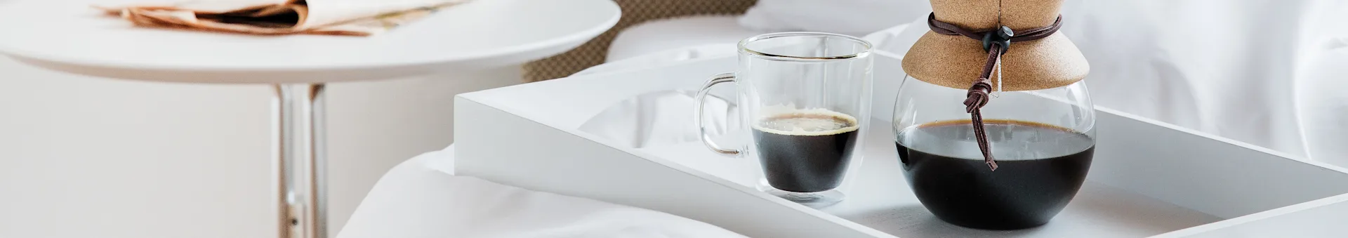 A white tray with a glass mug of coffee and a coffee carafe with a leather collar on a bed.