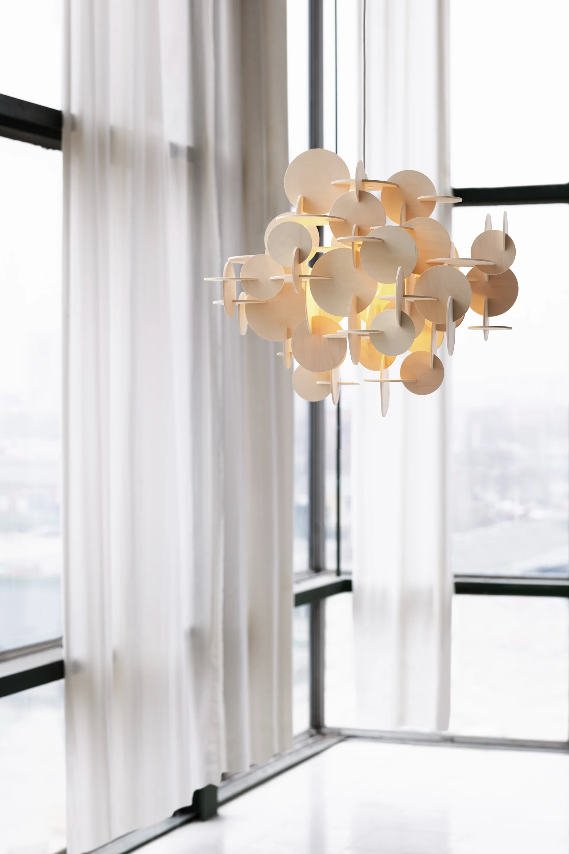 A unique wooden chandelier of stacked circular pieces, glowing warmly, hangs against large windows with white sheer curtains.