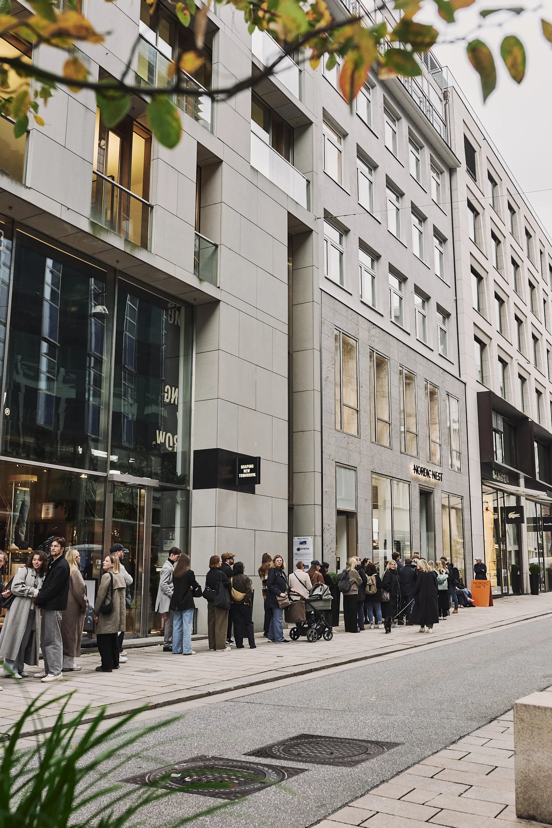 A long queue of people waiting outside a modern building with shopfronts, autumn leaves in the foreground.