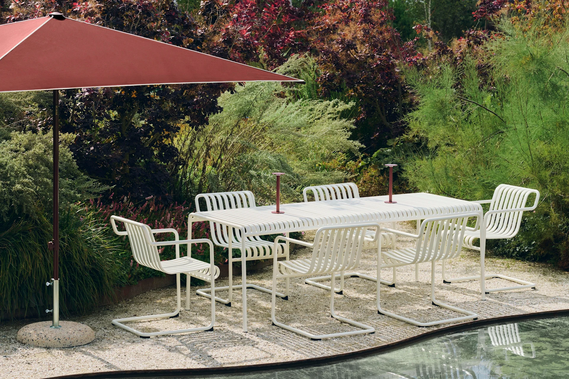 Outdoor dining set with a white slatted table and six chairs under a large red umbrella, on a gravel patio beside a pond.