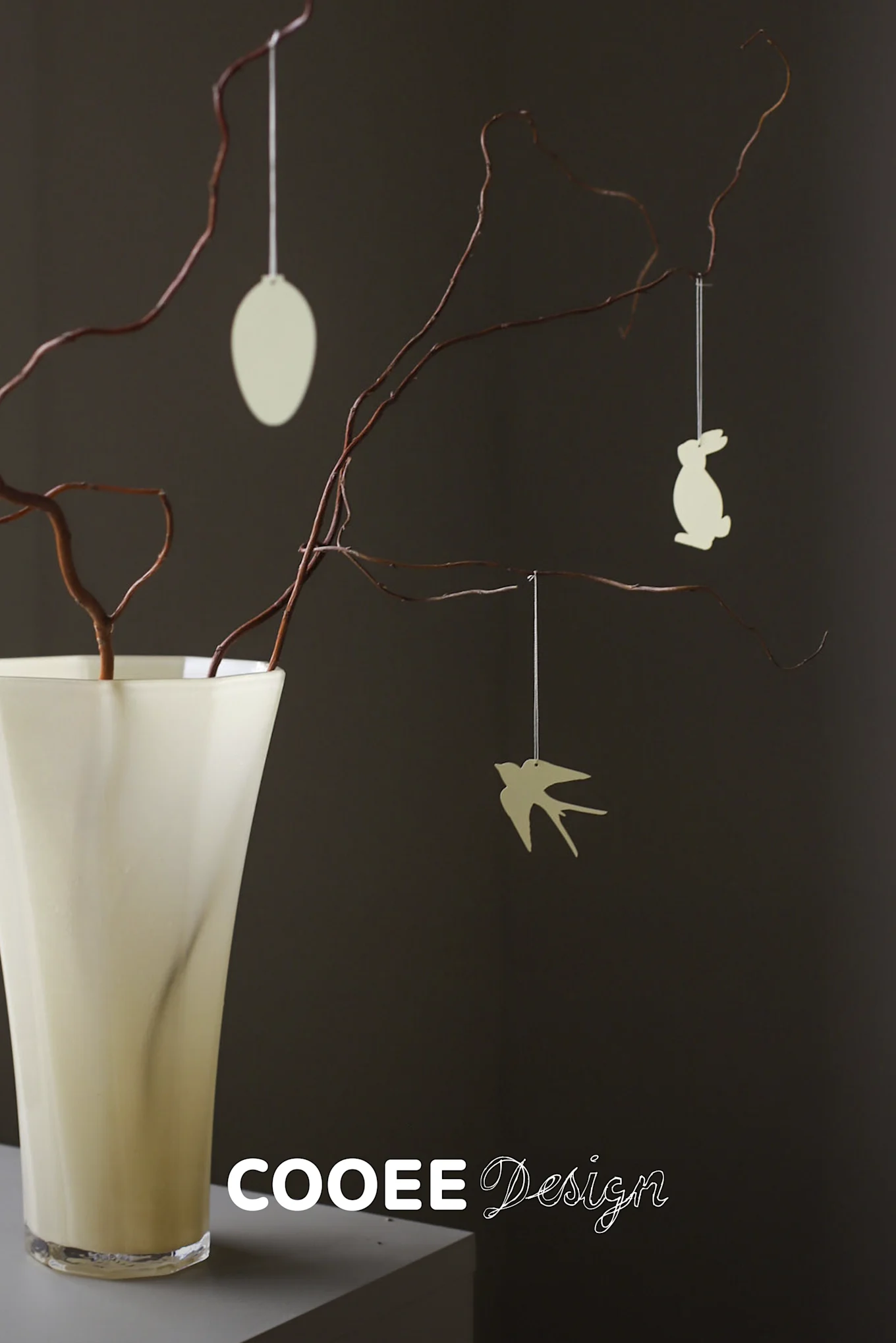Light-colored vase with dark branches, from which a cream egg, bunny, and bird ornament hang against a dark background.