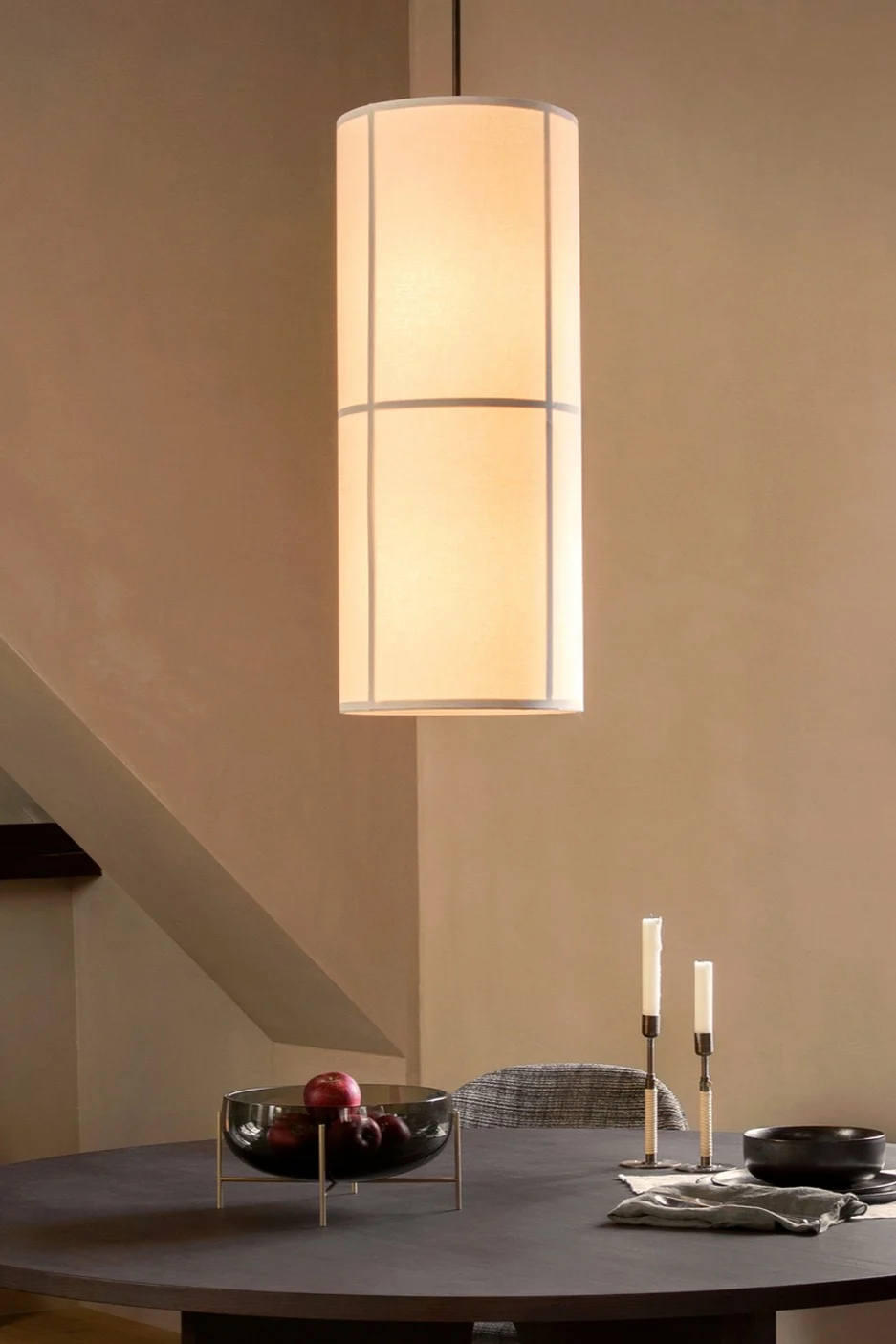 A cylindrical pendant light hangs over a dark dining table with a fruit bowl, candles, and a napkin.