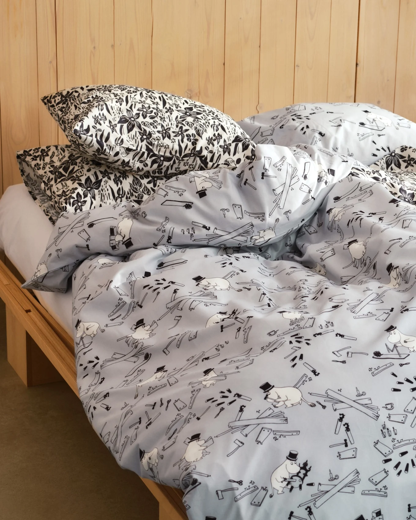Bed with pillows in black and white floral print and a light blue duvet with cartoon figures, tools, and wood planks. Wooden bed frame.