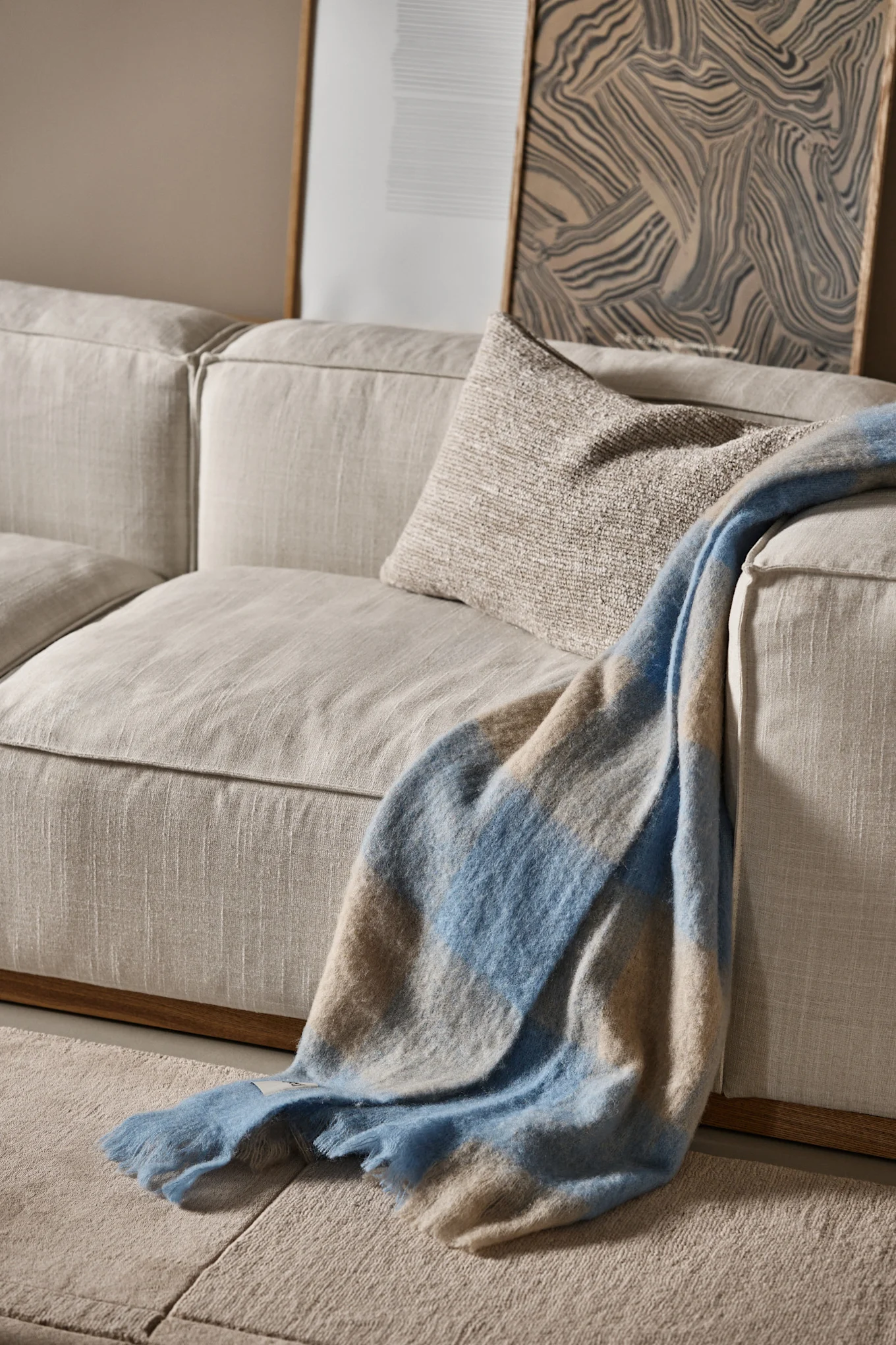 A light beige sofa with a textured beige cushion and a blue and beige plaid blanket draped over it. Abstract artwork is in the background.