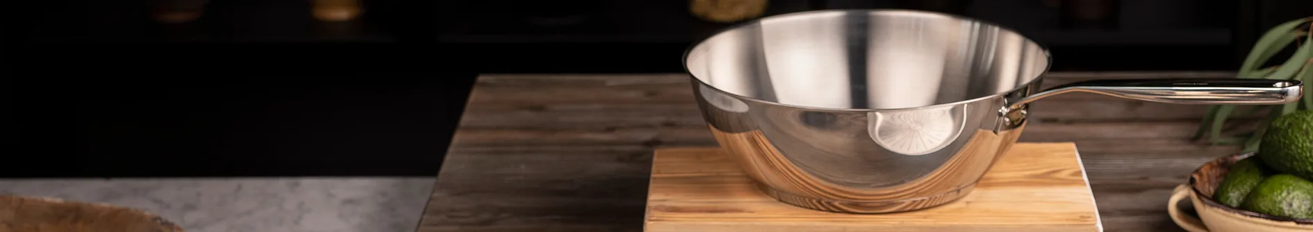 Shiny silver saucepan with a handle, sitting on a wooden cutting board, with a bowl of avocados visible.