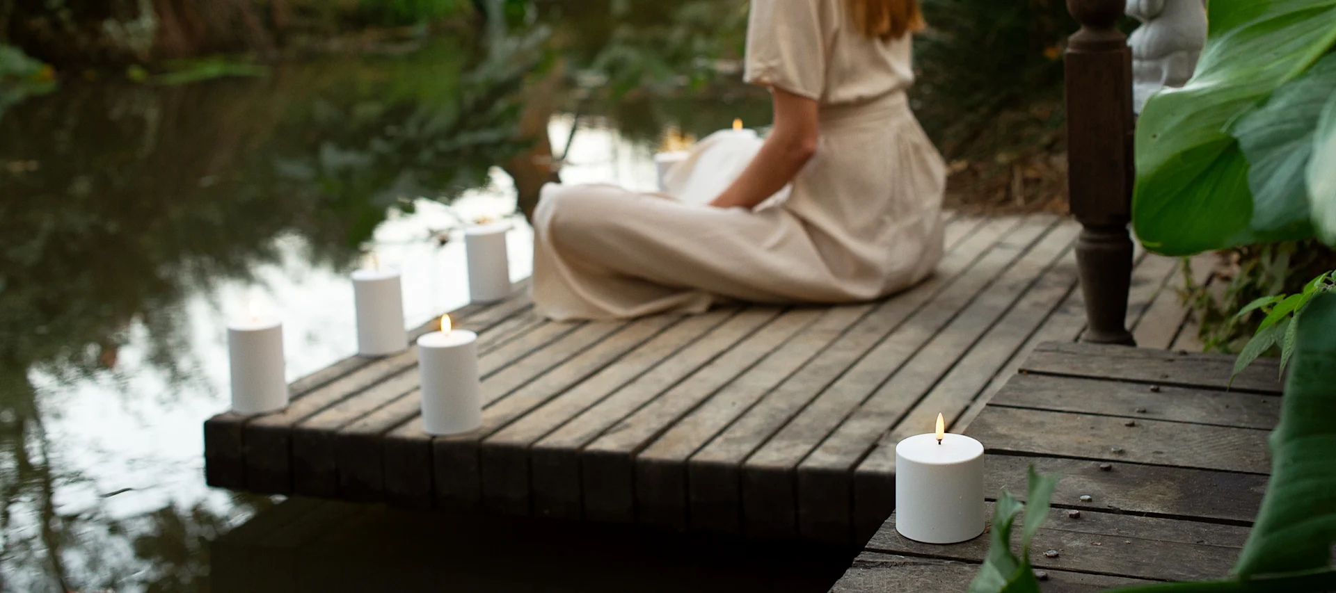 A person in light clothing sits on a wooden dock next to water, surrounded by several lit white pillar candles.
