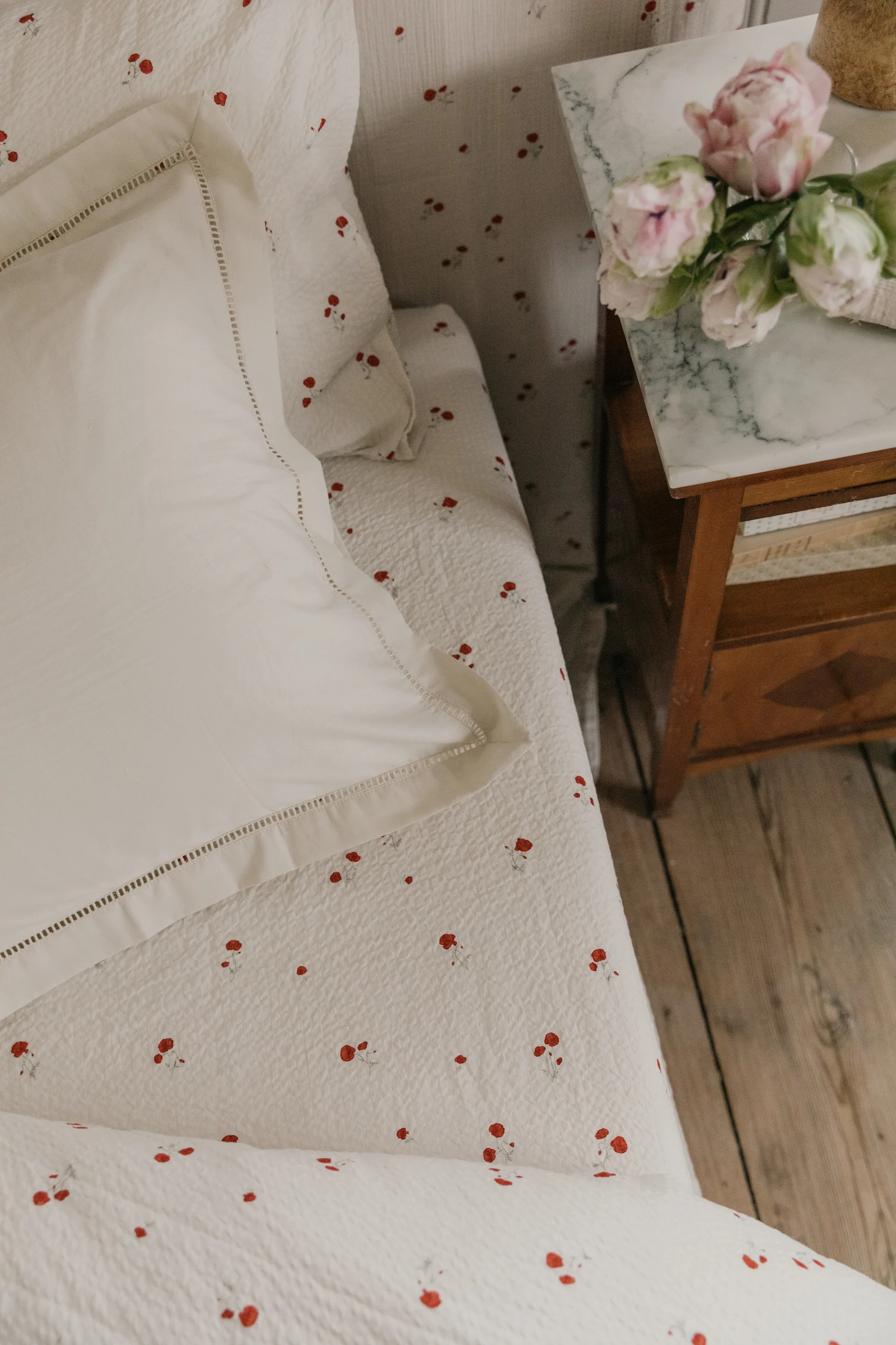 White bedding with a red floral pattern, a white pillow, and pink flowers on a marble bedside table.