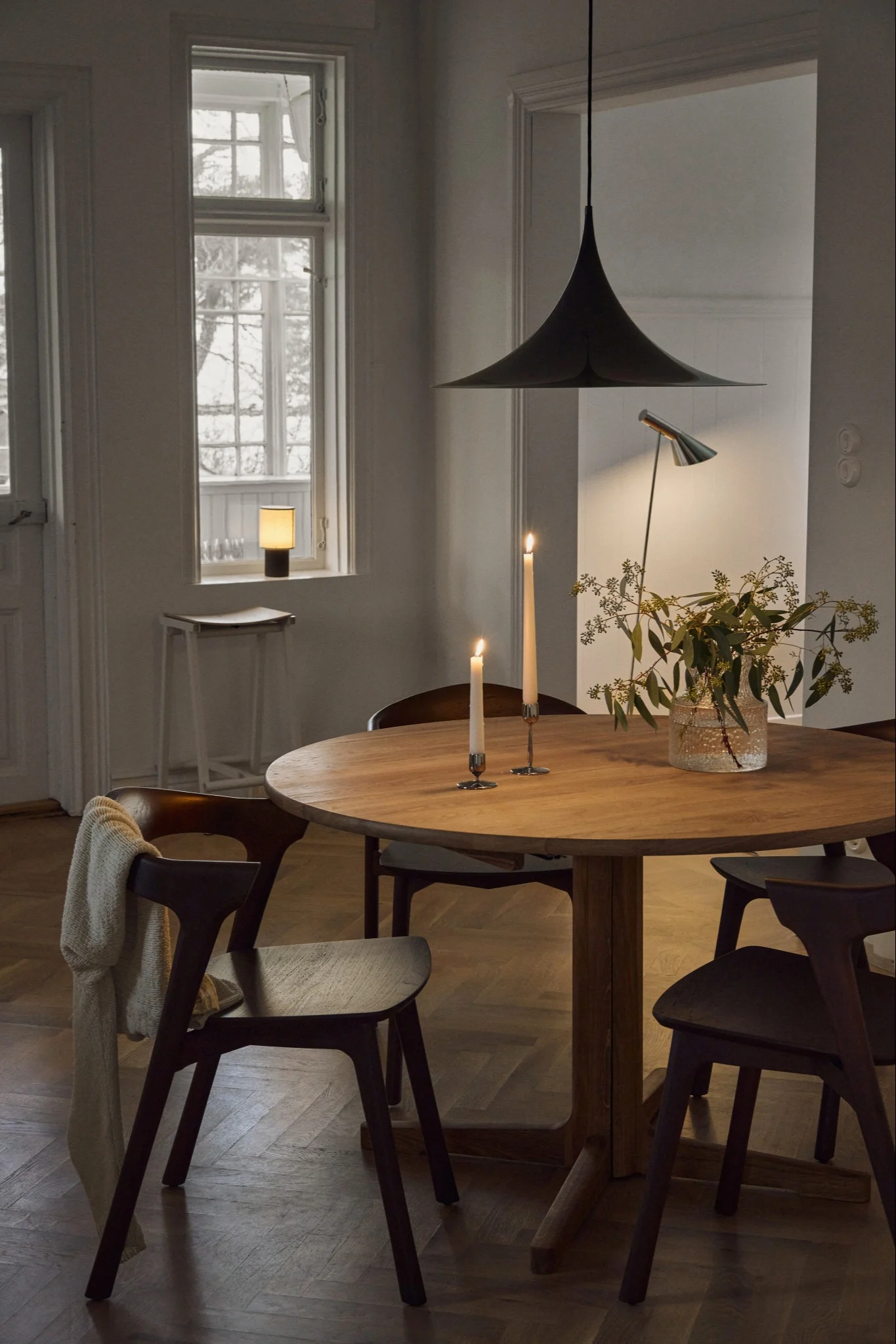 Cozy dining room with round wooden table, chairs, lit candles, foliage, and pendant lamp under a window.