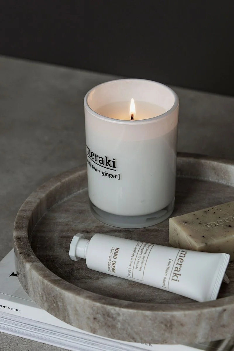 A lit 'Meraki' white tea and ginger candle, hand cream, and soap on a round marble tray.