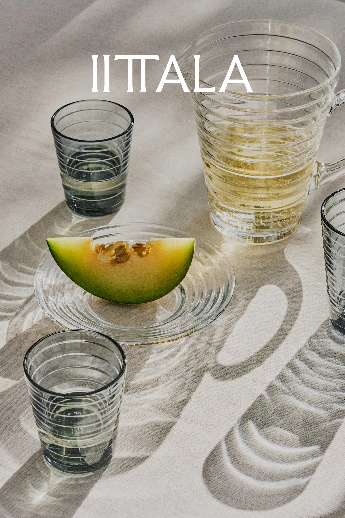 IITTALA logo above glass tumblers, a pitcher with sparkling drink, and a melon slice on a plate, all on a light fabric.