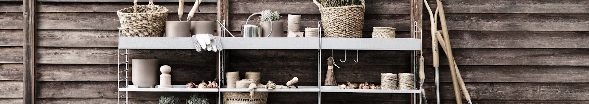 Rustic shelving unit against a wooden wall, displaying gardening tools, woven baskets, pots with plants, a watering can, and twine.