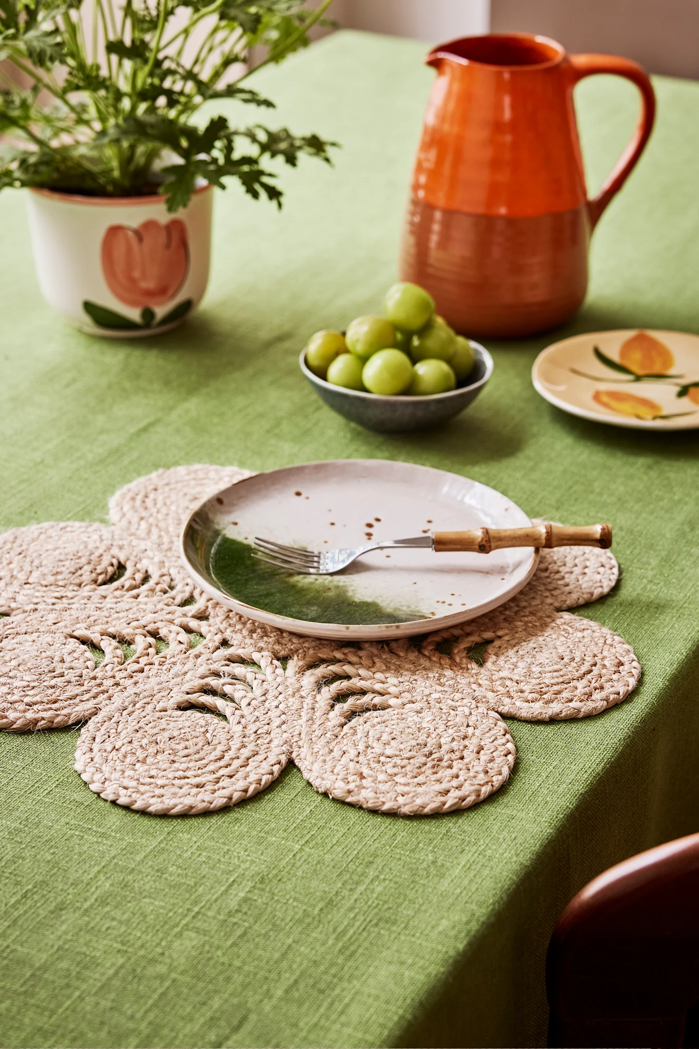 Table with green cloth, flower placemat, speckled ceramic plate, fork, potted plant, grapes, small plate, and orange pitcher.