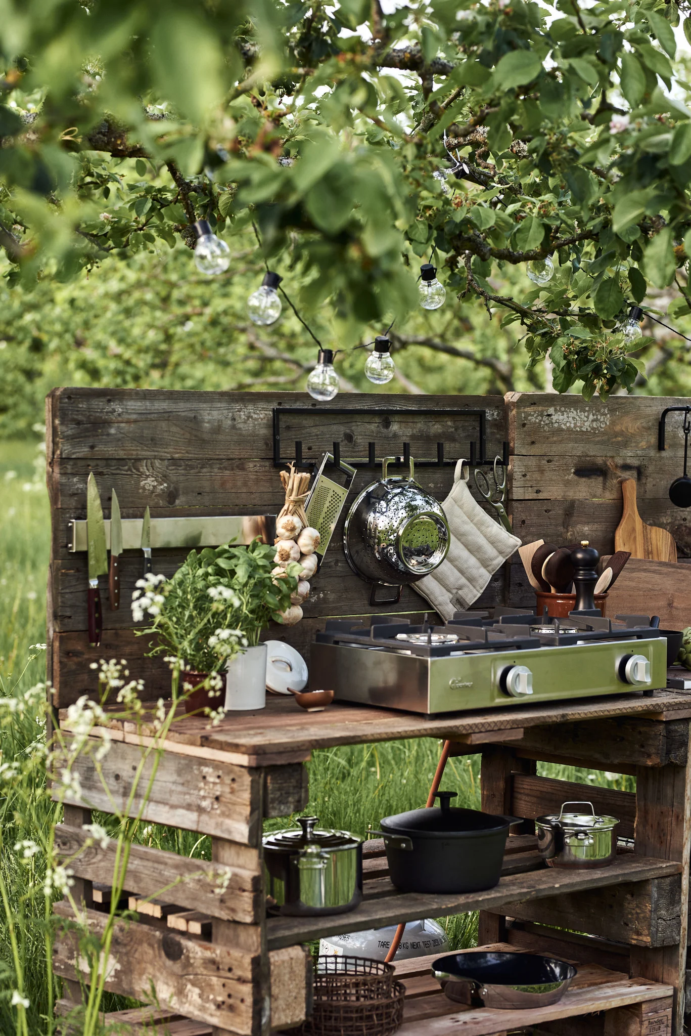 An outdoor kitchen built from wooden pallets, featuring a green two-burner gas stove, cooking utensils, herbs, and string lights.