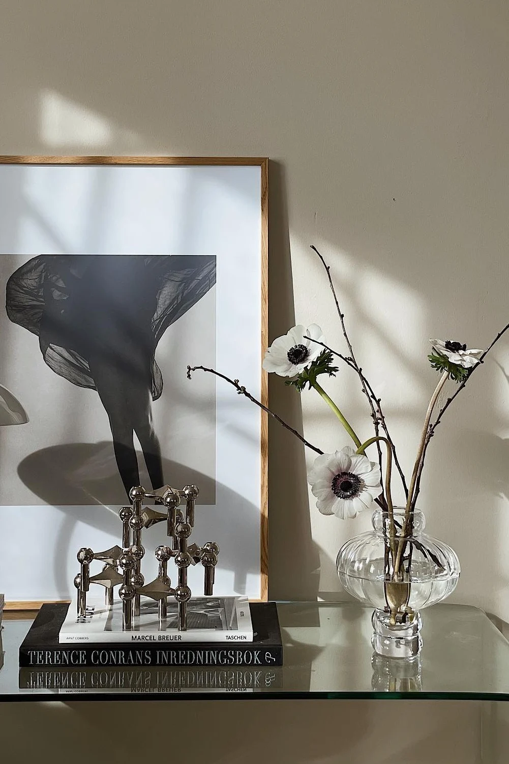 A framed black and white photo of legs with flowing fabric, a silver modular candle holder, books, and white anemones in a glass vase.