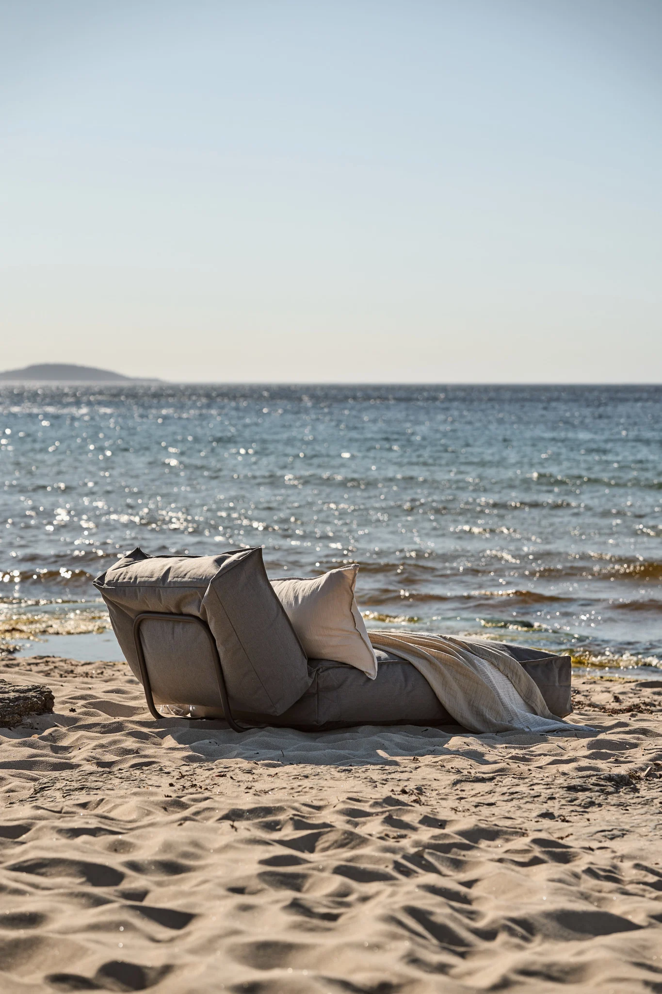 Grey lounge chair with off-white pillow and light blanket on a sandy beach by the sea.
