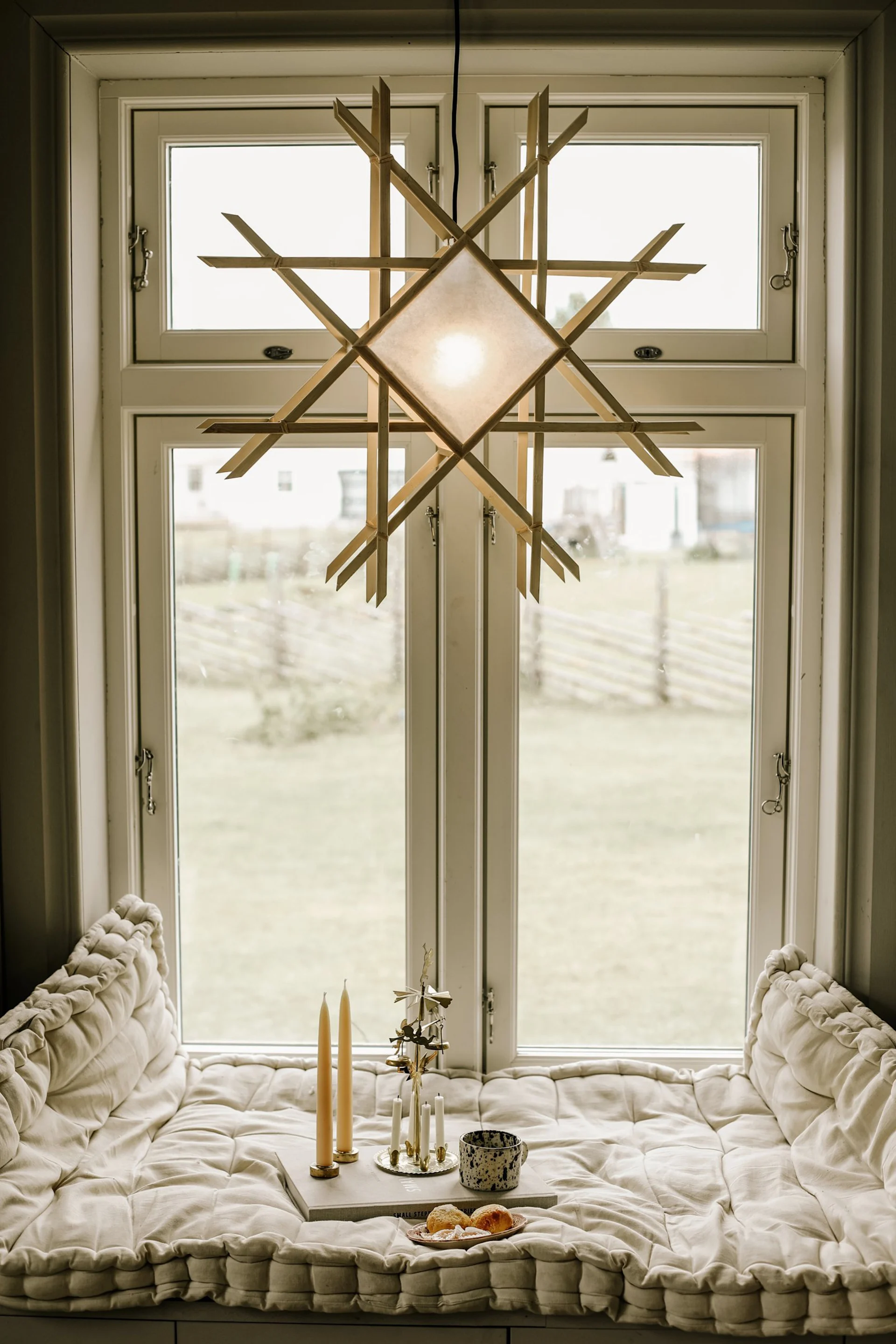 A star-shaped light fixture hangs in front of a window, above a tufted window seat with candles, a mug, and pastries.