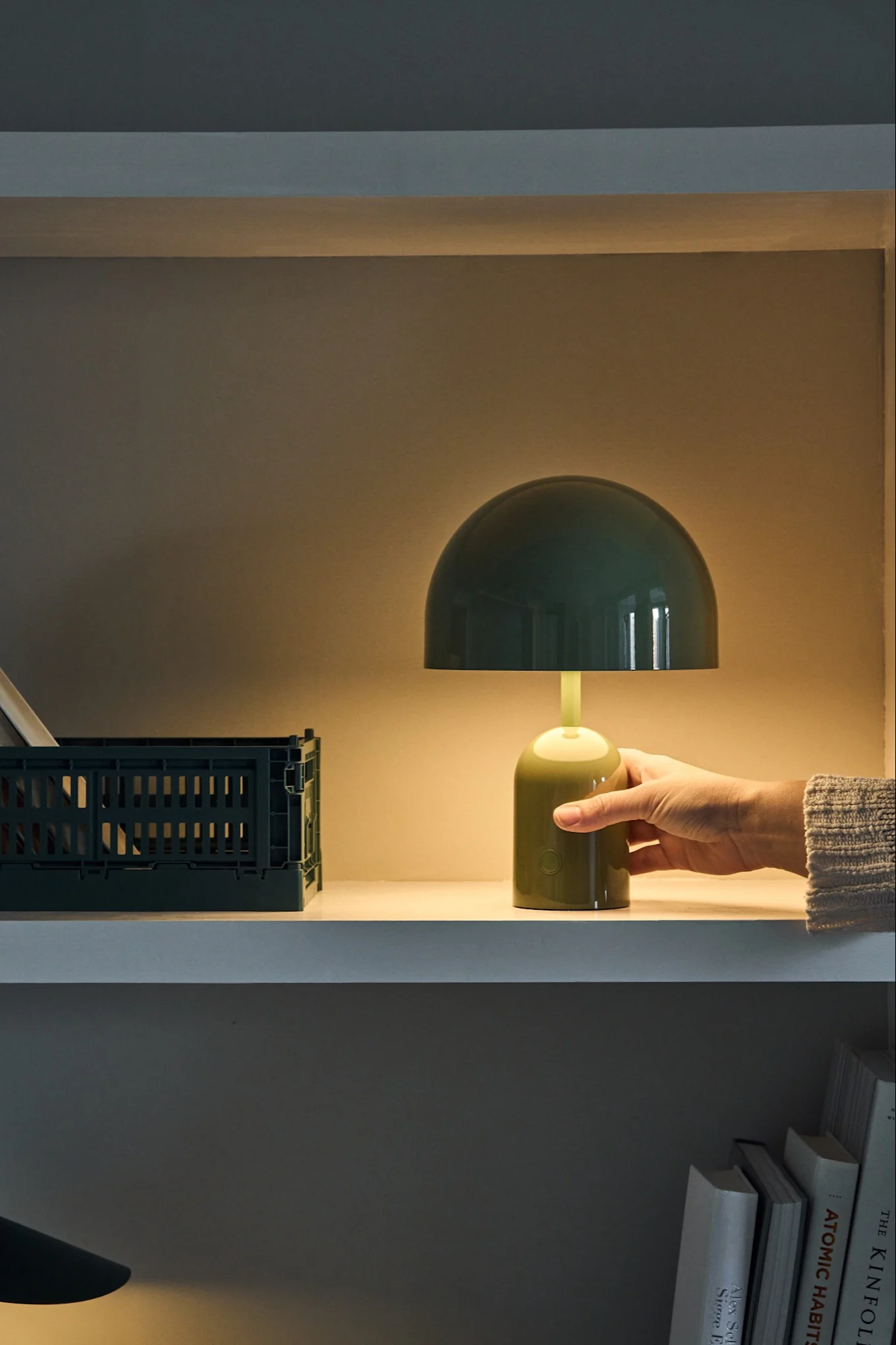 A hand adjusts a lit, green mushroom-shaped table lamp on a shelf next to a dark green crate.