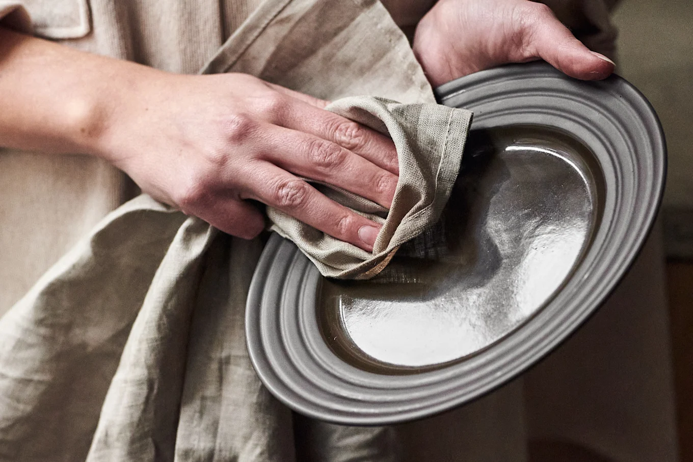 Hands drying a dark gray ceramic plate with a light beige linen cloth.