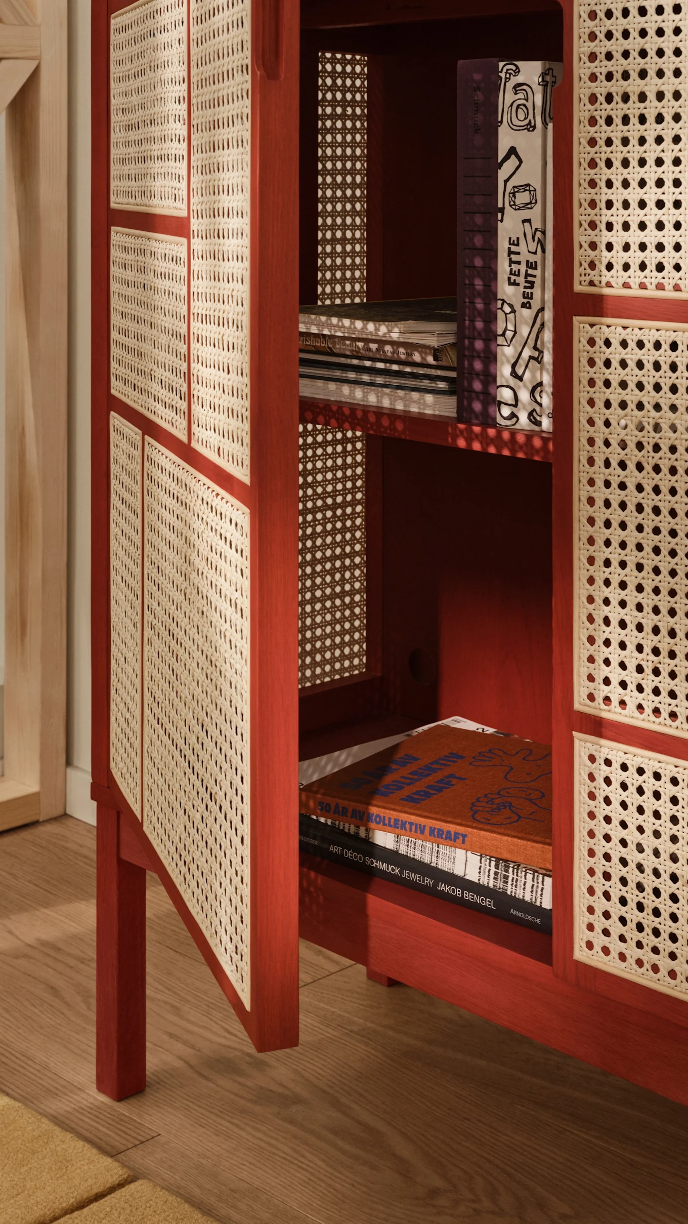 Red wooden cabinet with cane webbing doors, one slightly open showing books, on a wooden floor with a yellow rug.