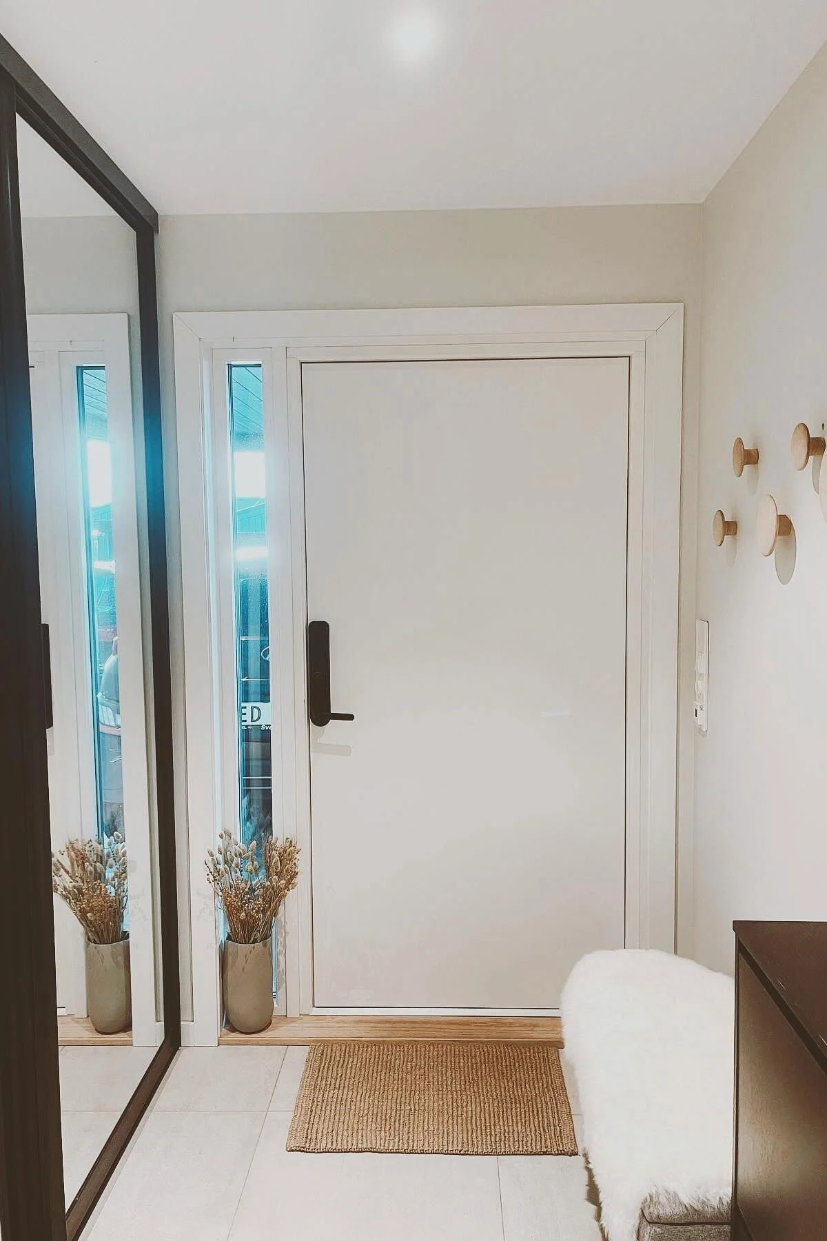 Bright entryway with white door, natural doormat, dried flowers, mirror, wooden wall hooks, and a fluffy white bench.