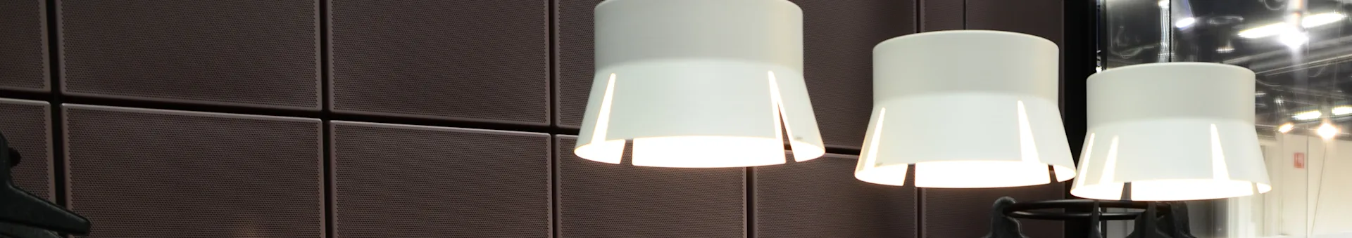 Three white conical pendant lights hanging in front of a dark brown, perforated panel wall.