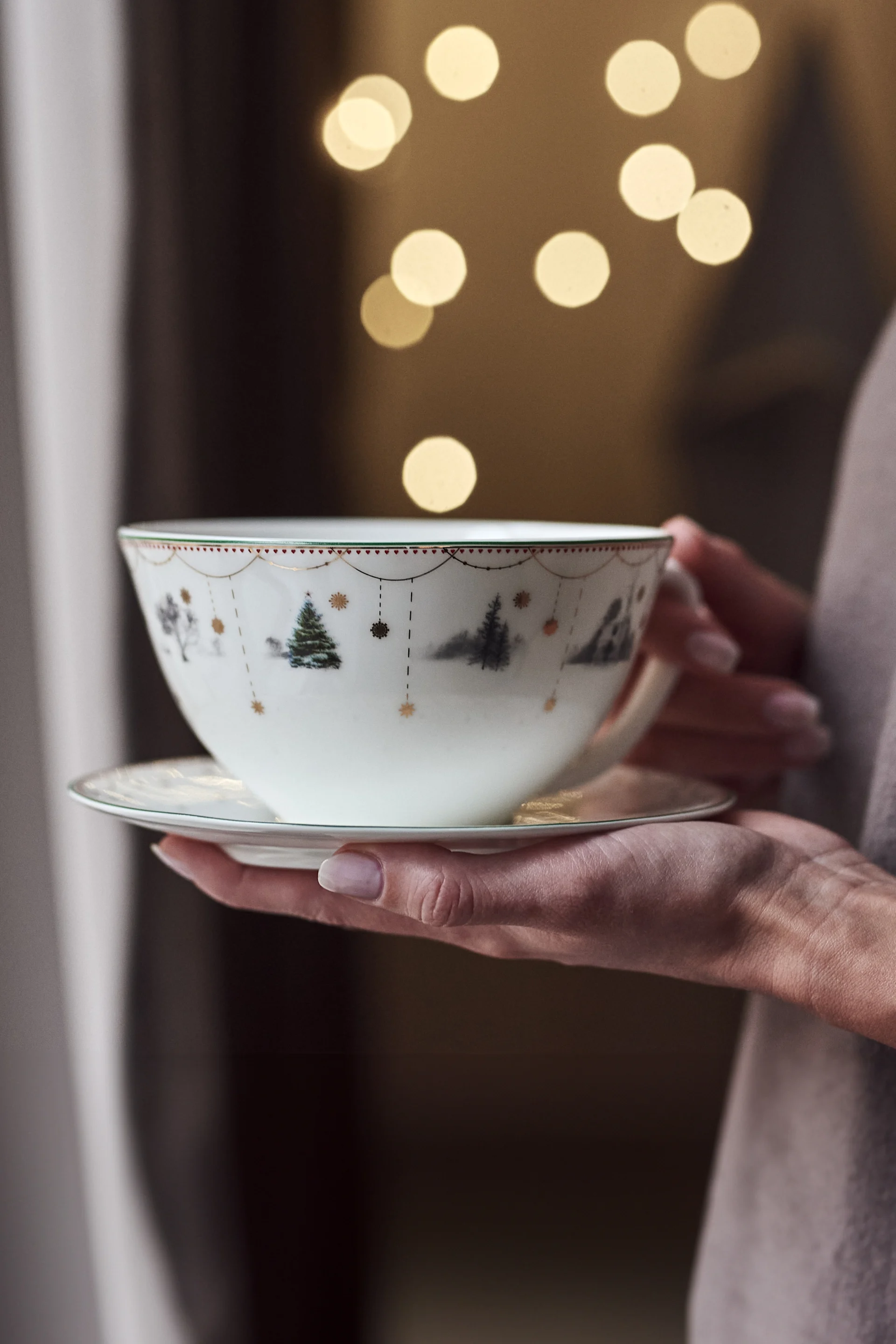 Hands hold a white teacup and saucer with a festive winter scene, evergreen trees, and red hearts, against warm bokeh lights.