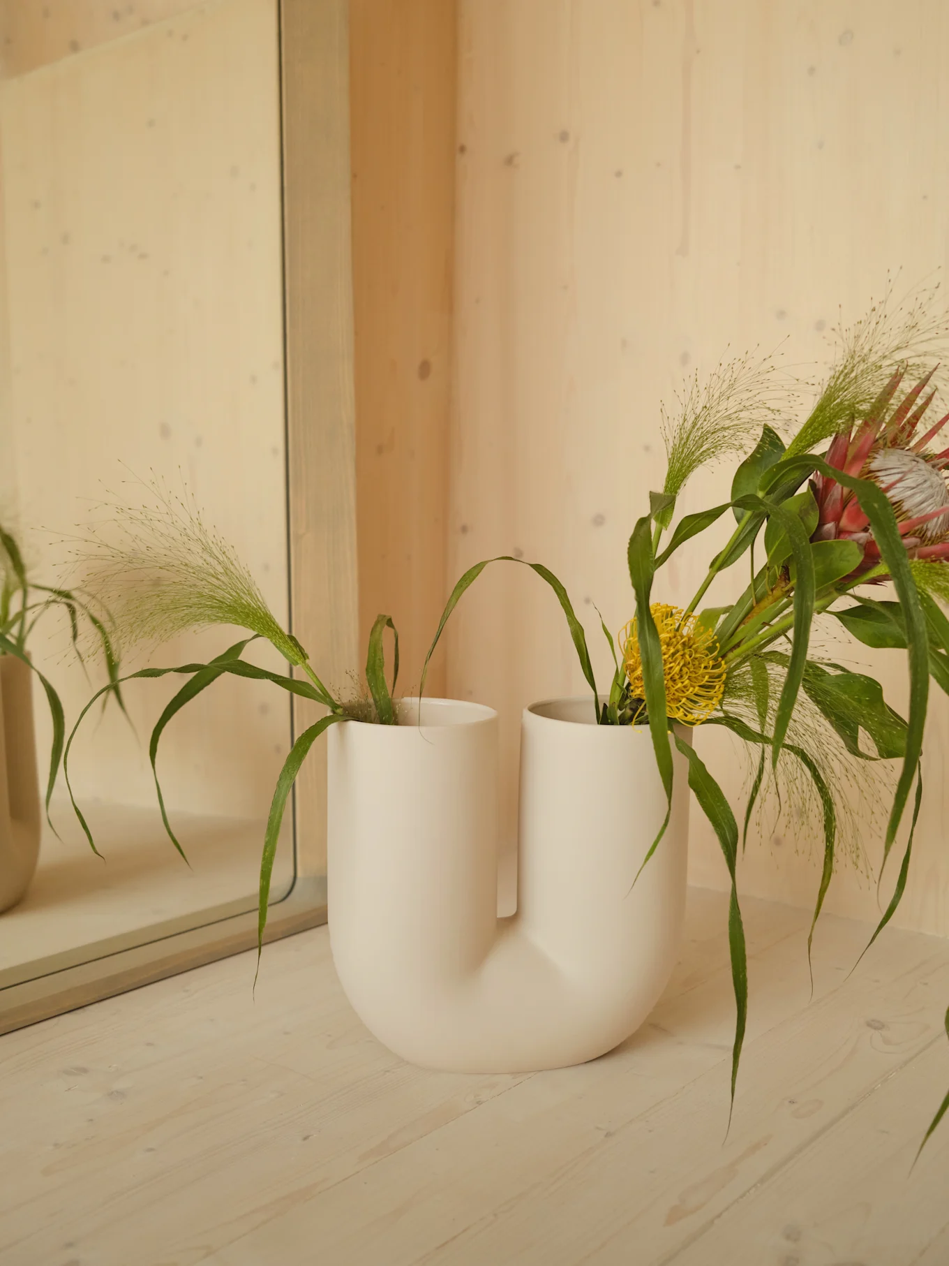 U-shaped white vase holds pink protea, yellow flower, and green foliage on a light wood surface.
