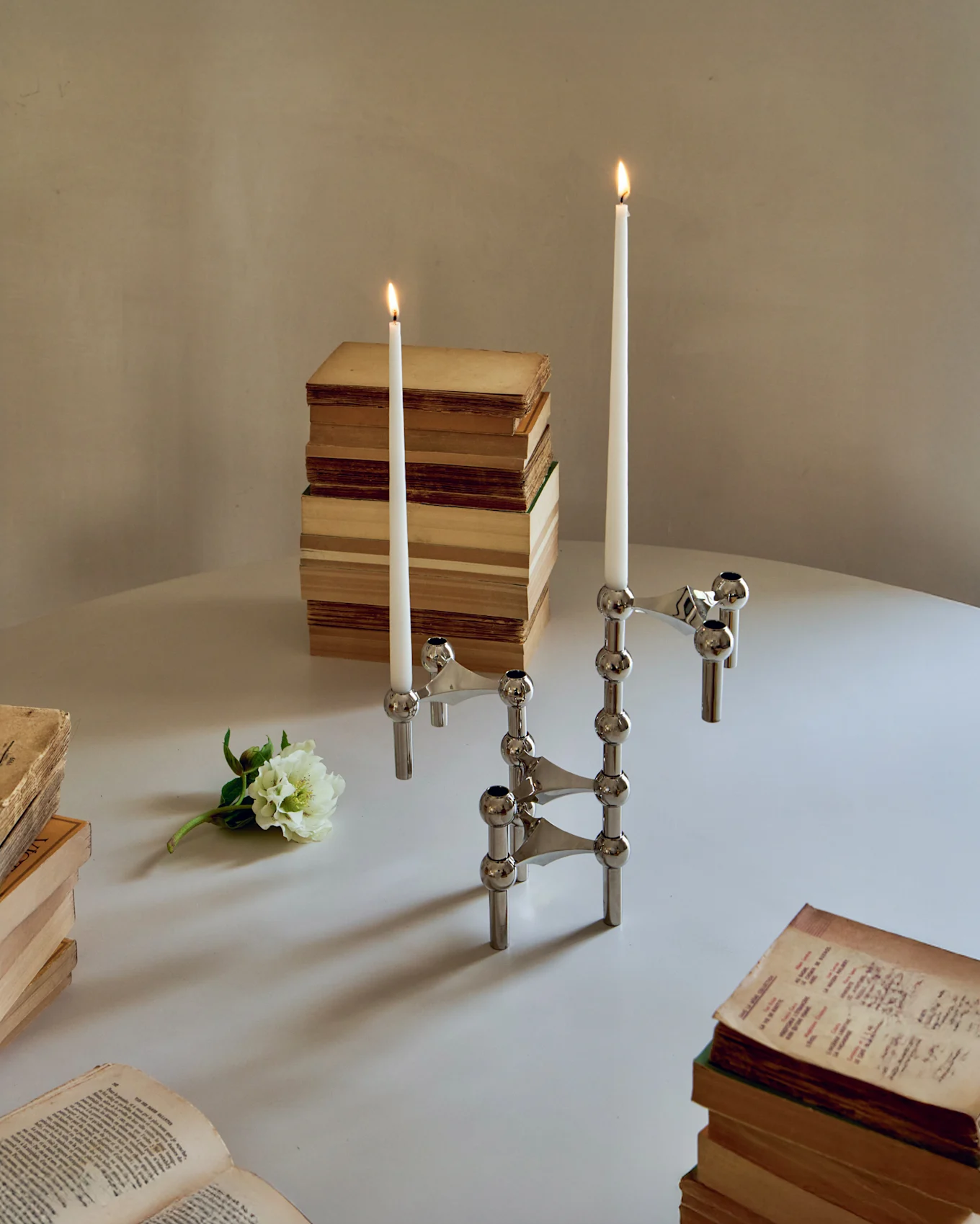 Two chrome modular candle holders with lit white taper candles, surrounded by old books and a white flower on a table.