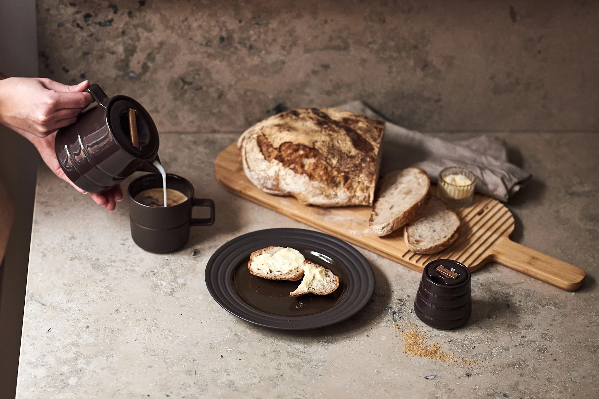 Hand pouring milk into a dark mug of coffee, with buttered bread slices, a rustic loaf, and sugar on a countertop.