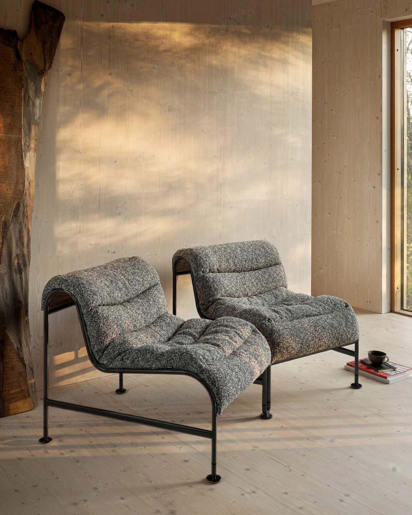 Two modern low-profile lounge chairs with grey textured upholstery and black metal frames sit on a light wood floor in a sunlit room.