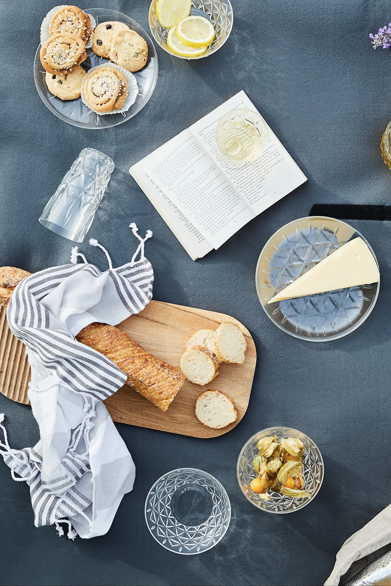 Overhead view of a picnic with cookies, lemons, wine, cheese, baguette, physalis, and an open book on a grey blanket.