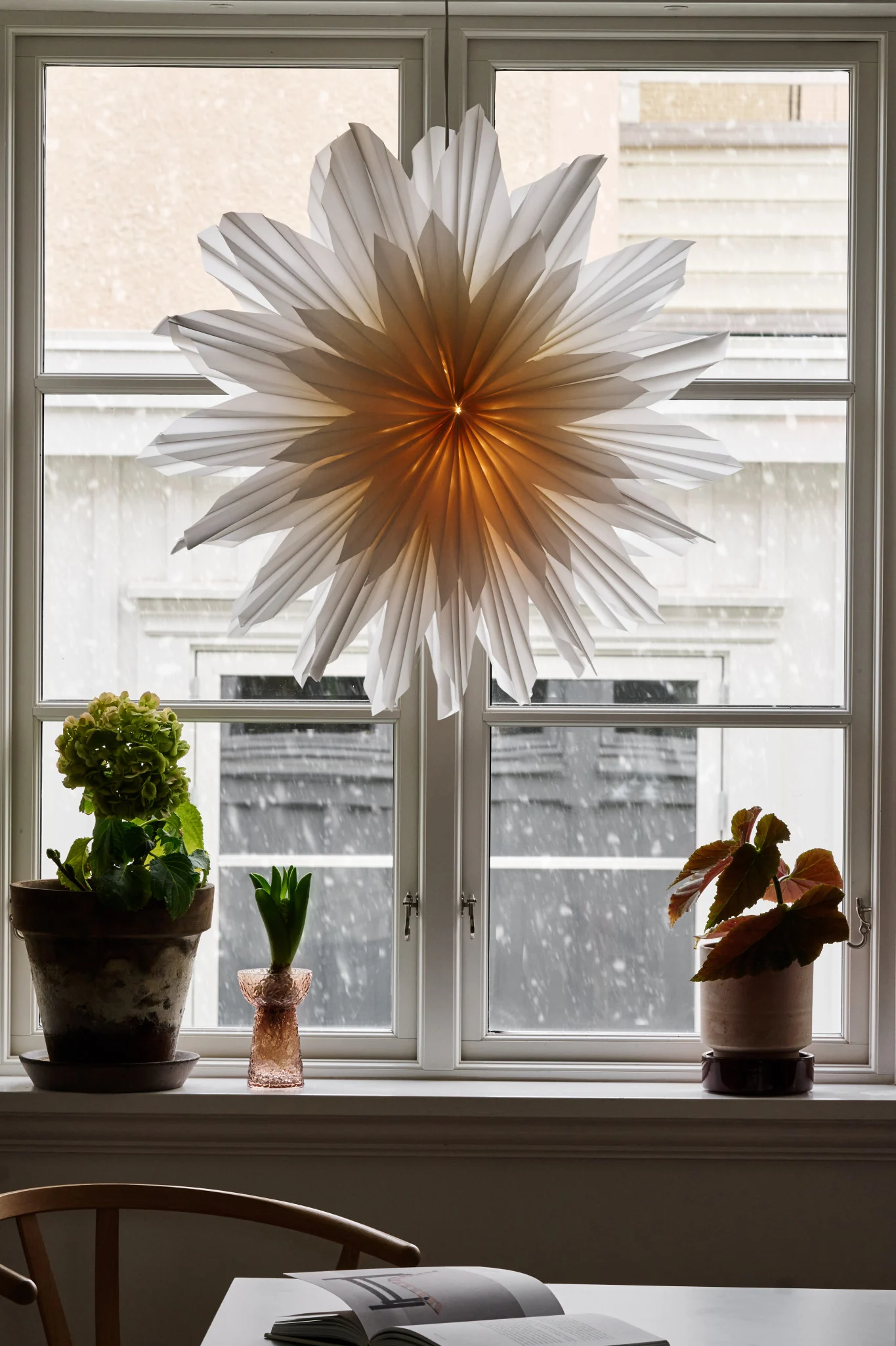 White pleated paper star hanging in a window, with snow falling outside and potted plants on the windowsill.