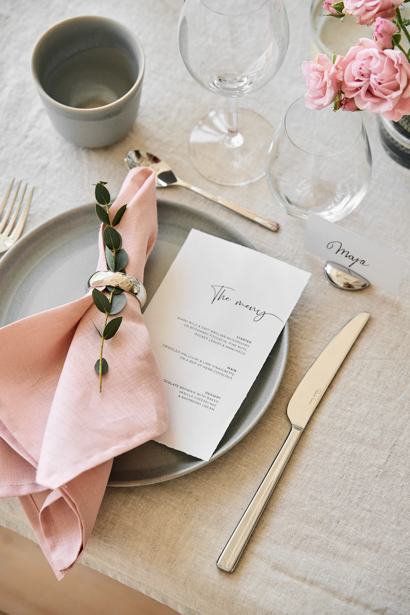 Elegant dining table set with a grey plate, pink linen napkin, green foliage, menu, and name card.