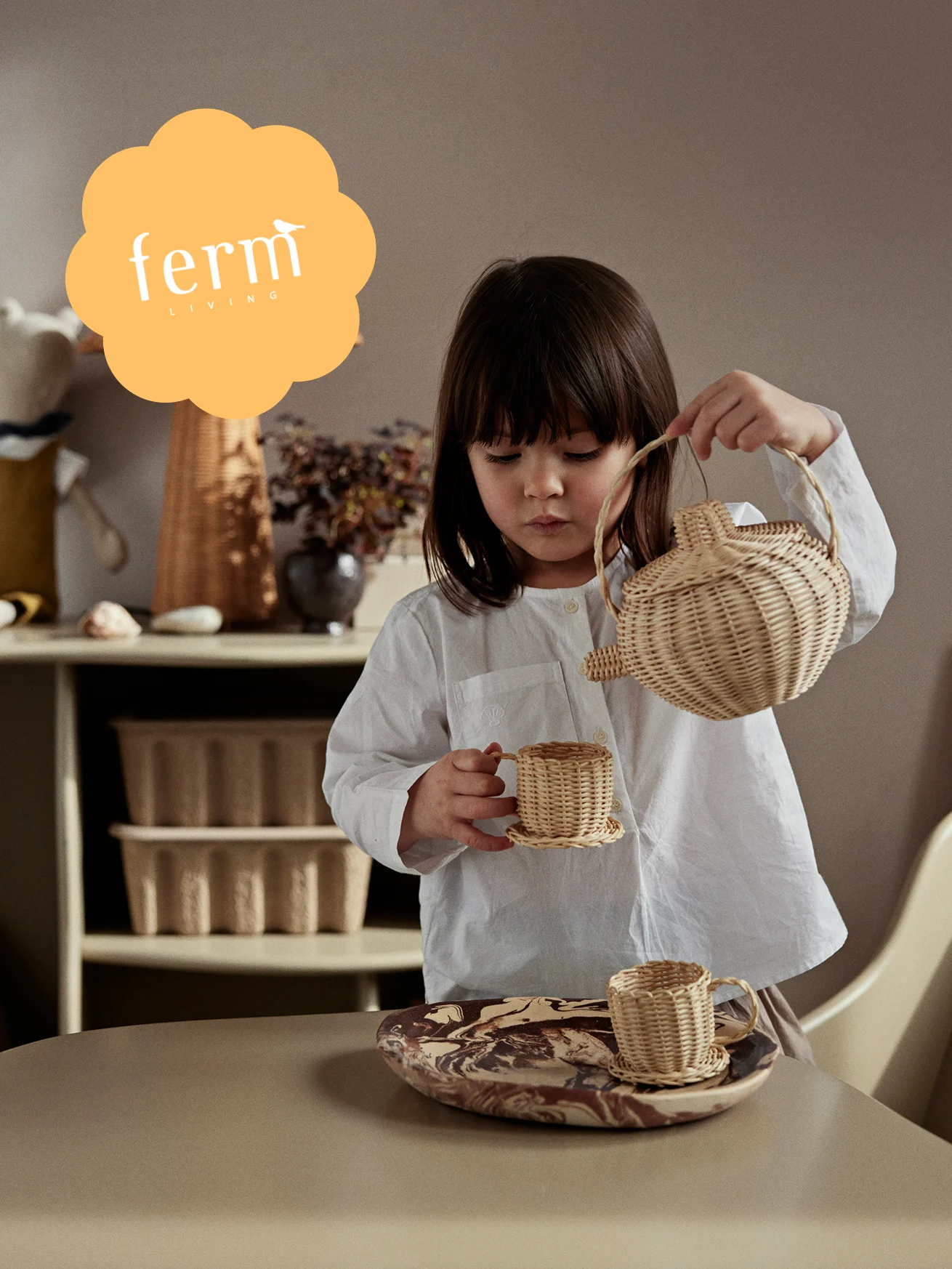 Child in white shirt plays with a woven wicker tea set, pouring into a small cup. Ferm LIVING logo in orange cloud.