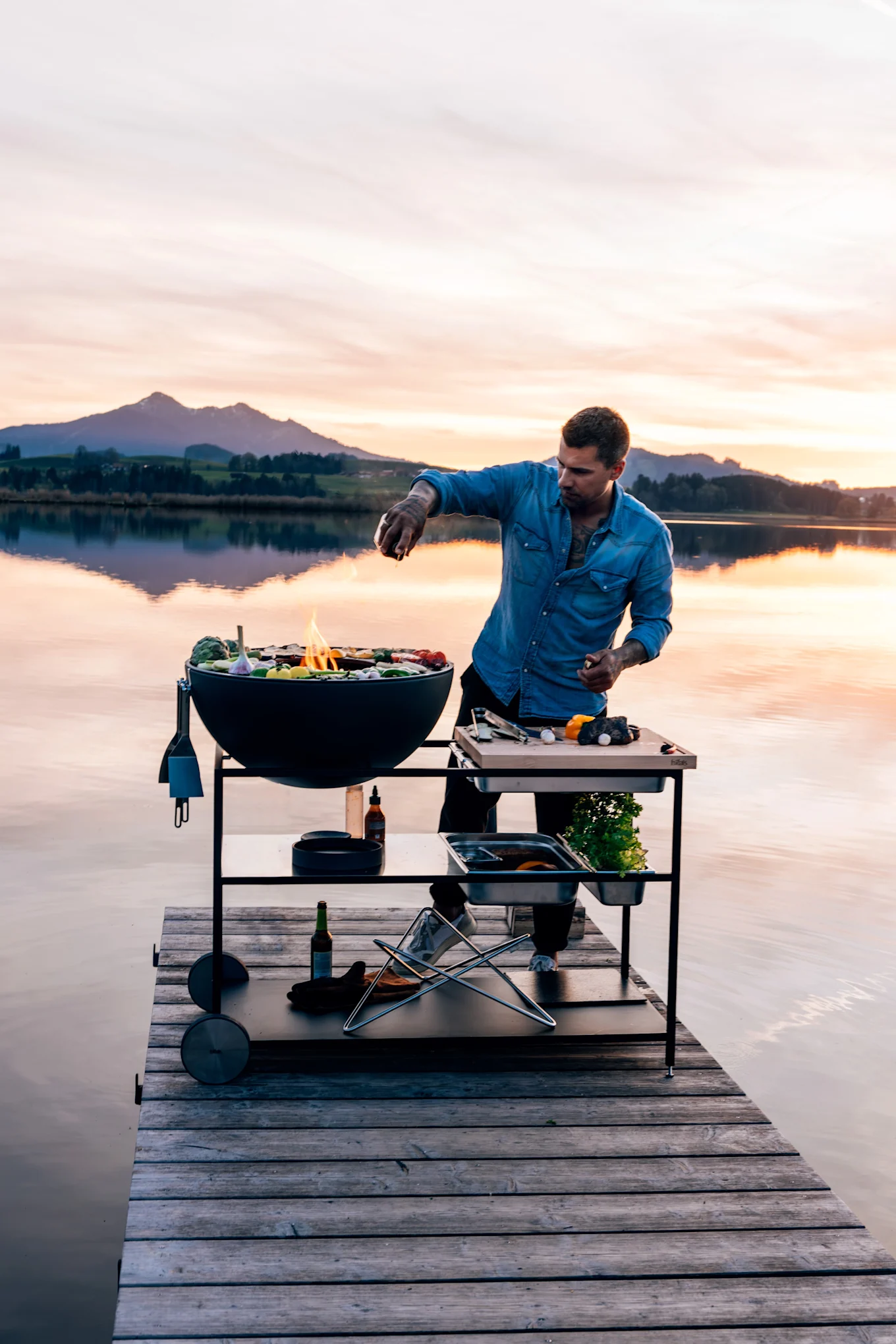 Man in a denim shirt grilling food on a round black grill and preparation table on a wooden pier at sunset.