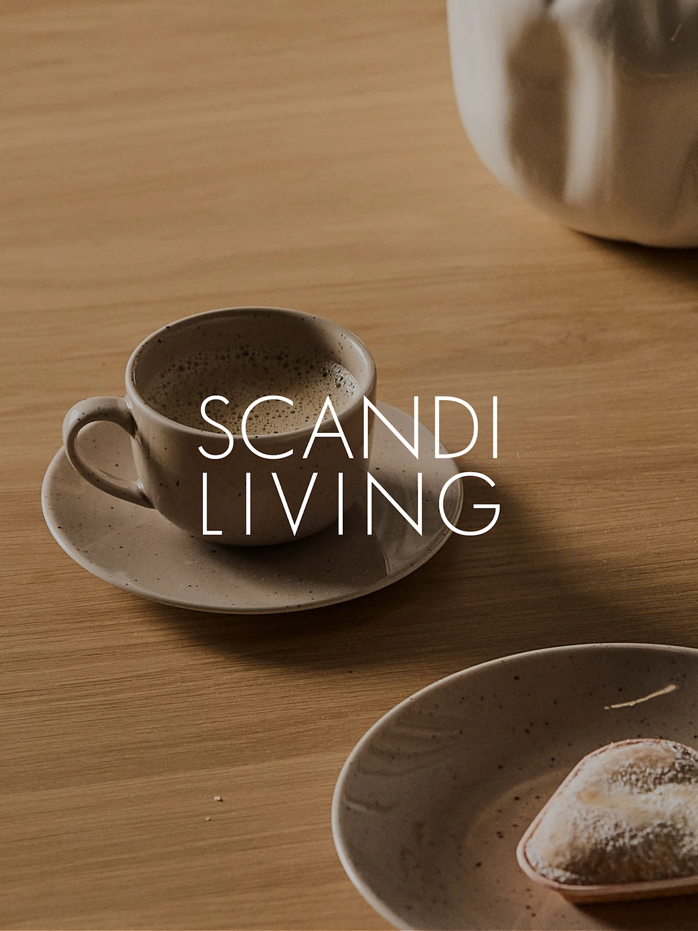 A ceramic cup of coffee on a saucer with a pastry on a wooden table, featuring the text 'SCANDI LIVING'.