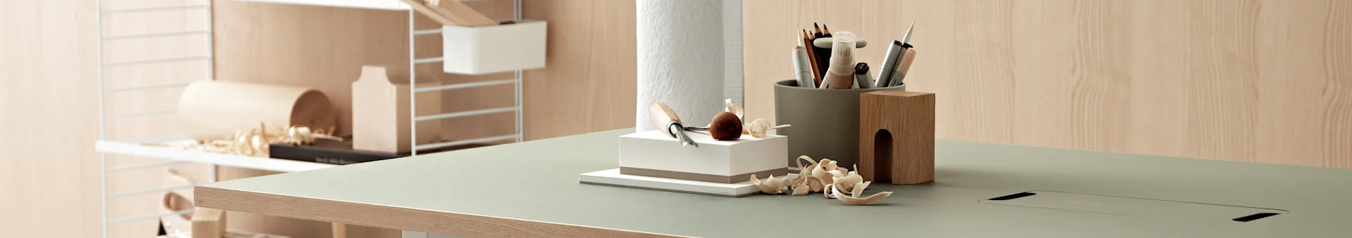 A light green desk with wood edge, featuring various crafting tools, pencils, and wood shavings on its surface. A wooden shelf unit is in the background.