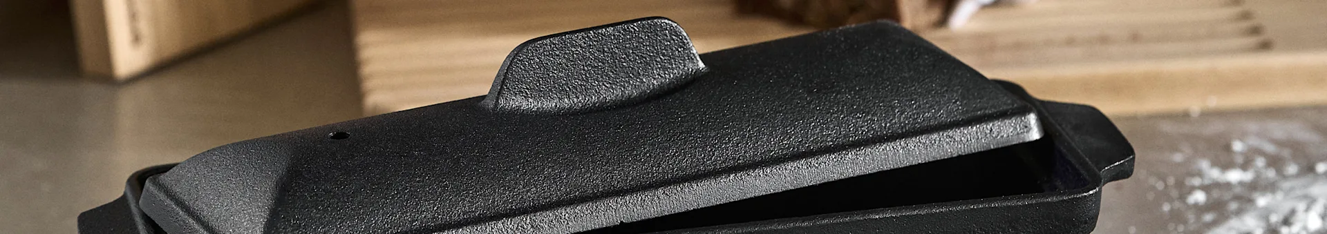 Close-up of a black cast iron loaf pan with a textured finish and its lid slightly ajar, on a blurred background.