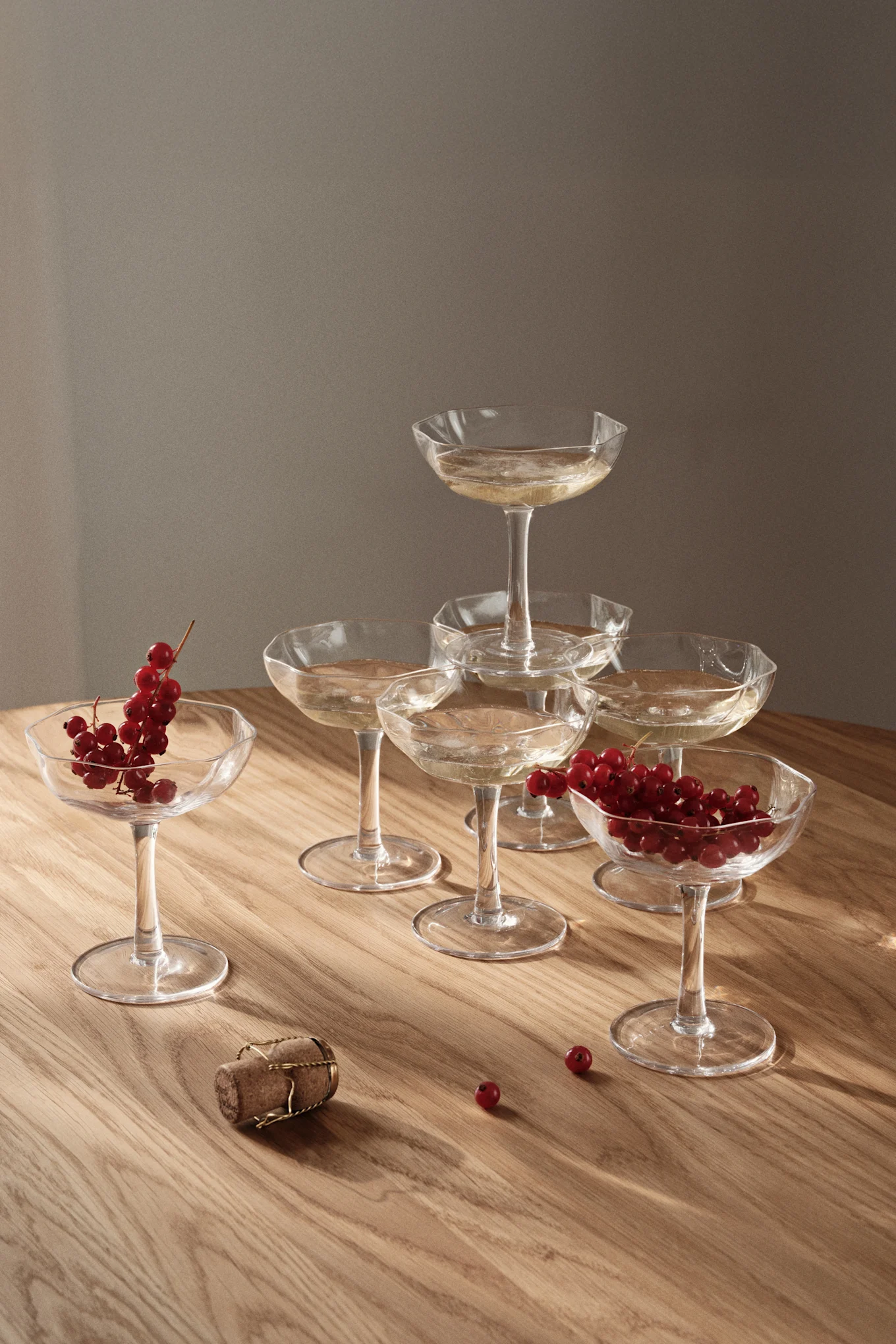 Champagne coupe glasses stacked in a pyramid, filled with sparkling wine, alongside loose red currants and a cork on a wooden table.