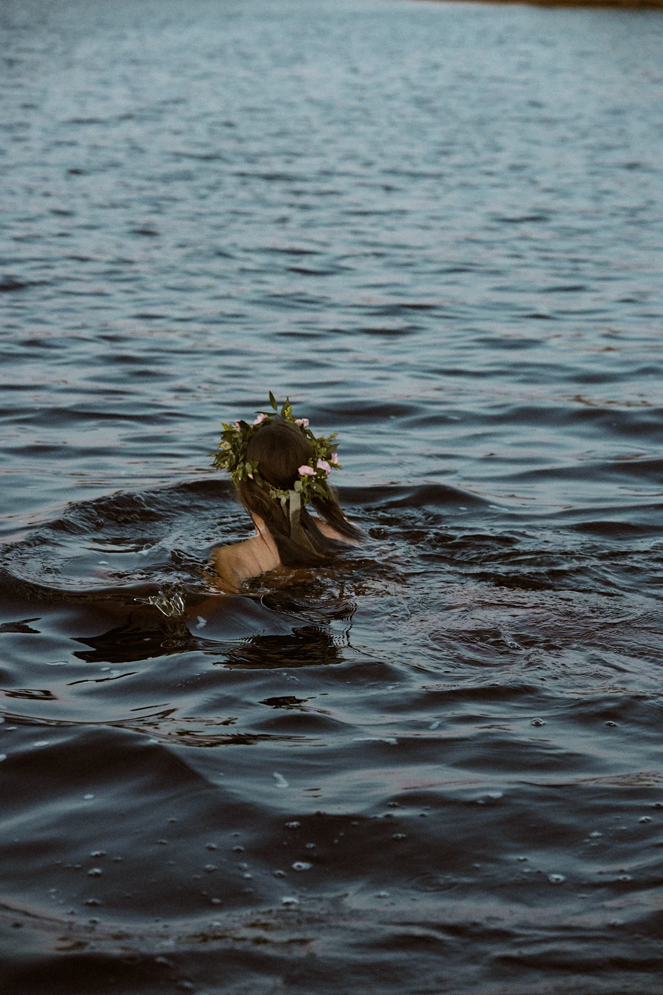 Person with a flower crown swimming in dark water, seen from behind.