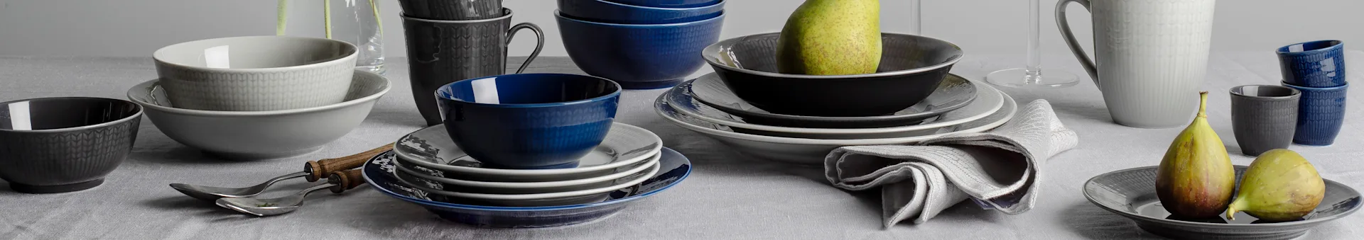 Assortment of blue, grey, and black ceramic bowls, plates, and mugs with textured patterns on a grey tablecloth, with pears and figs.