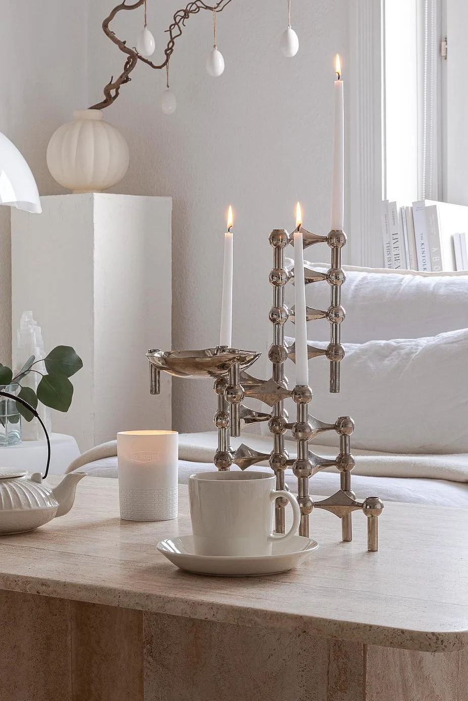 Modern silver modular candle holder with three lit white candles, a teacup, and teapot on a stone table with Easter decor.