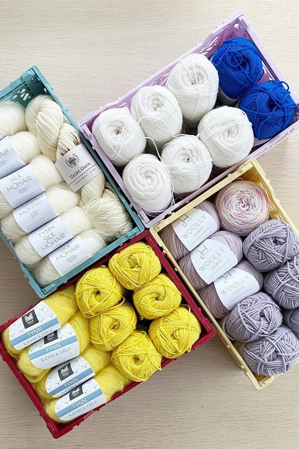 Overhead view of four crates filled with many balls of yarn in cream, white, blue, yellow, and purple on a light wooden surface.