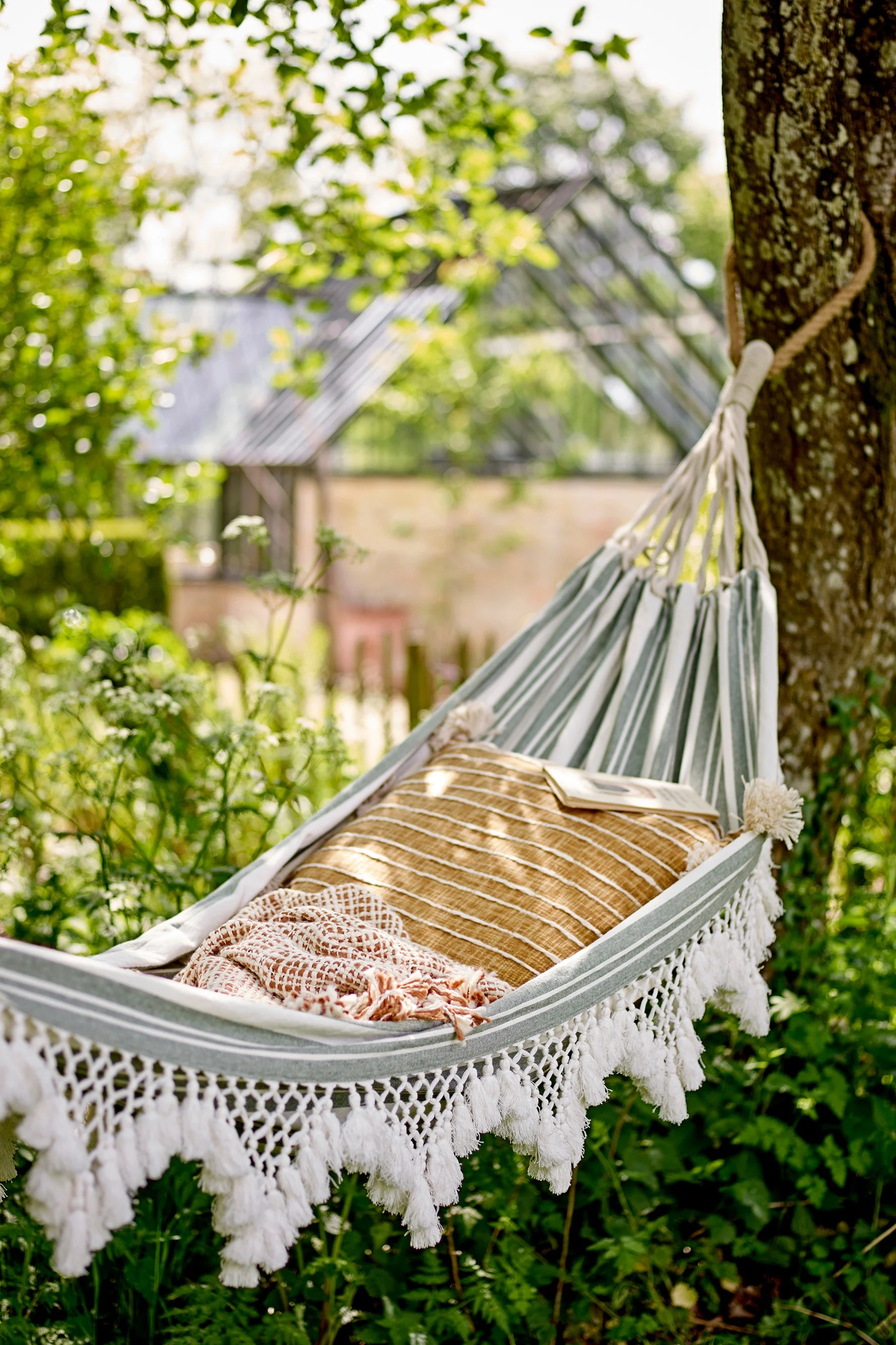 A striped hammock with a pillow and blanket hangs in a lush green garden with a greenhouse in the background.